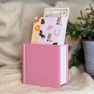 May include: A pink square note holder containing a stack of white paper sheets. Several cards are tucked into the holder, including one with a pattern of brown horses, pink cowboy hats, and tan boots, and another with a floral design in orange and pink.