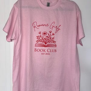 May include: Pink t-shirt with a red graphic design of an open book with flowers and the text "Romance Girly Book Club Est 2024"