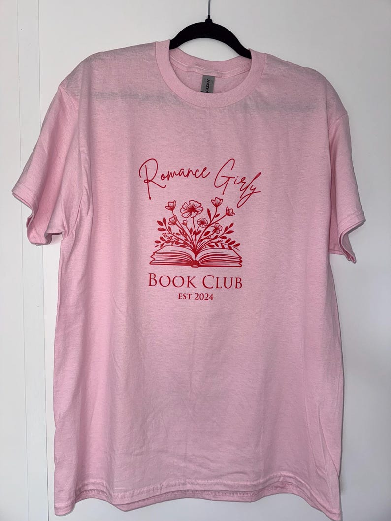 May include: Pink t-shirt with a red graphic design of an open book with flowers and the text "Romance Girly Book Club Est 2024"