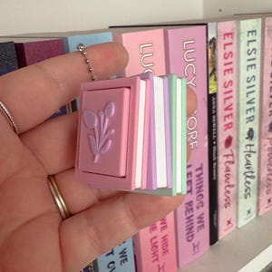 May include: A small, pink book-shaped charm with a floral design, attached to a chain. The charm has a stack of colourful pages. In the background, a bookshelf displays several books with pink and pastel covers.