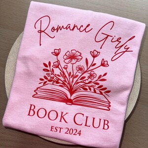 May include: Pink t-shirt with red text that says "Romance Girly Book Club Est 2024". The text is surrounded by a red illustration of a book with flowers.