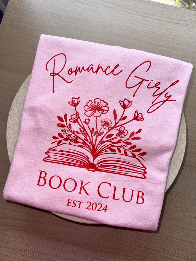 May include: Pink t-shirt with red text that says "Romance Girly Book Club Est 2024". The text is surrounded by a red illustration of a book with flowers.