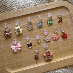 May include: A selection of decorative earrings featuring charms like teddy bears, books, flowers, and a gingerbread man. The earrings are presented on a wooden tray against a neutral backdrop.