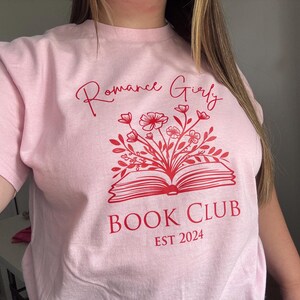 May include: Pink t-shirt with the text "Romance Girly Book Club Est 2024" in red. The design features a red open book with flowers growing out of it.