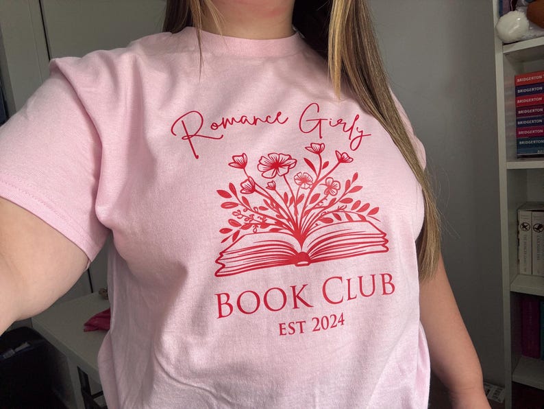May include: Pink t-shirt with the text "Romance Girly Book Club Est 2024" in red. The design features a red open book with flowers growing out of it.
