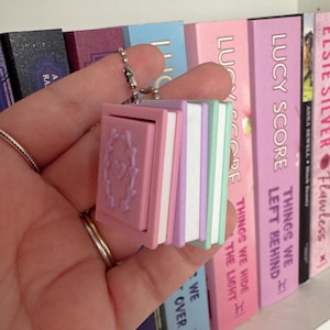 May include: A small book-shaped charm with a pink cover and a heart design, attached to a silver chain. The charm has a stack of pastel-coloured pages in pink, purple, and green. The charm is held in front of a bookshelf.