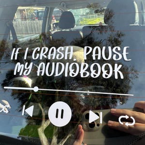 May include: White vinyl decal on a car window, featuring the text "If I crash, pause my audiobook" and a media player design. The image includes a person's hand holding a phone.