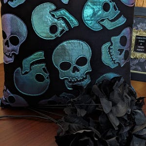 Peut inclure: Coussin carré noir avec des appliques de crânes bleu-vert irisés. Les crânes sont de différentes tailles et orientations. Des roses noires sont au premier plan. Une image encadrée avec le texte "A BEAUTIFUL THING IS NEVER PERFECT" est en arrière-plan.