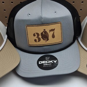 May include: Three trucker hats with a leather patch. One hat is tan with a white mesh back and a patch that says "WYO". The other two hats are tan and gray with the number "307" on the patch. Each hat has a "Decky Classic Original" sticker.