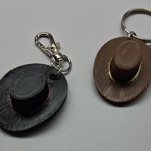 May include: Two cowboy hat keychains. One is black with a red trim, attached to a silver clip. The other is brown with a yellow trim, attached to a silver keyring. Both have a textured design.