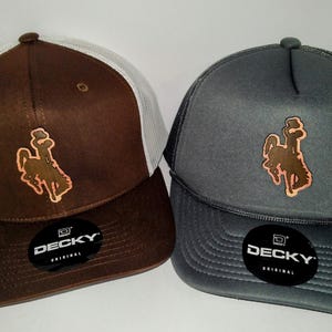 May include: Two trucker hats with a brown leather patch featuring a silhouette of a cowboy riding a bucking horse. The hats are brown and gray with a white mesh back. The hats have the word "DECKY" printed on the front.