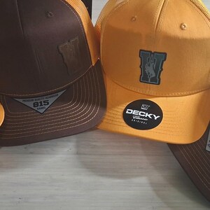 May include: Four baseball caps with a trucker hat design. The caps have a yellow front panel and a brown or black top. Each cap has a patch with a design and the brand name "DECKY" on a sticker.