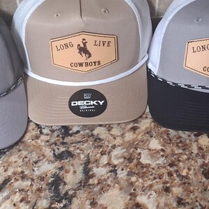 May include: A collection of five trucker hats with a "Long Live Cowboys" patch. The hats feature various color combinations, including gray, black, and tan, with mesh backs and curved brims. The patch shows a cowboy riding a horse.