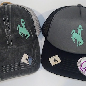 May include: Two trucker hats with a distressed denim look. One hat is black with a gray mesh back and the other is gray with a black mesh back. Both hats feature a green embroidered design of a cowboy riding a horse.
