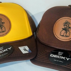 May include: Two trucker hats with brown mesh backs. One hat is yellow with a brown leather patch featuring a cartoon cowboy riding a horse. The other hat is brown with a brown leather patch featuring a cartoon cowboy riding a horse. Both hats have the word "DECKY" in white letters on a black tag.