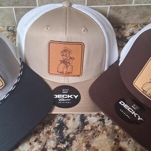 May include: Three trucker hats with a cowboy graphic on a square leather patch. The hats have different color combinations: gray and black, tan and white, and brown and white. Each hat has a mesh back and a curved brim.