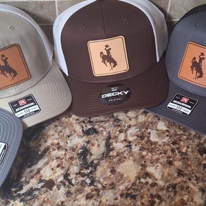 May include: Five trucker hats in various colors: gray, brown, and tan. Each hat features a square leather patch with a silhouette of a person riding a horse. The hats have a mesh back and a curved brim.