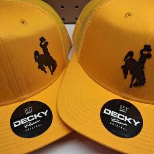 May include: Two yellow trucker hats with a brown silhouette of a person riding a horse. The hats have a mesh back and a curved brim. Each hat has a black and white sticker that says "DECKY Classic Original".