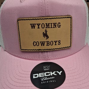 May include: Pink and white trucker hat with "WYOMING COWBOYS" patch. The patch is brown with a cowboy graphic. The hat has a curved brim and a mesh back. A black sticker with "DECKY Classic Original" is on the brim.