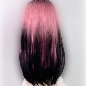 May include: A long, straight wig with a pink to black ombre colour gradient. The wig is styled with a centre parting and falls to the shoulders.