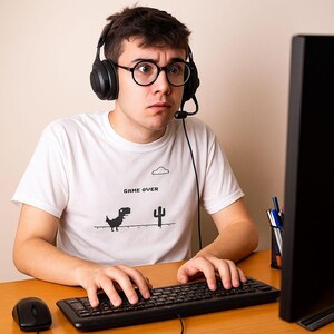 May include: White t-shirt with a black graphic of a pixelated dinosaur and cactus, with the words "GAME OVER". The person is wearing black headphones and glasses, and is sitting at a desk with a computer.