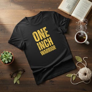 May include: Black t-shirt featuring the text "ONE INCH WARRIOR" in a yellow, distressed font. The shirt is on a wooden surface, with a succulent, a cup of tea, and an open book visible in the background.