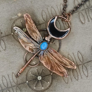 May include: A copper dragonfly pendant necklace featuring a blue gemstone and a black crescent moon charm. The dragonfly has detailed wings and hangs from a chain. This unique necklace is a piece of jewellery.