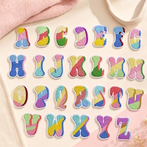 Op de afbeelding: Een complete set van zesentwintig geborduurde alfabetletter-patches, elk voorzien van een veelkleurig, diagonaal gestreept patroon in tinten roze, geel, blauw, groen en paars met een dikke witte rand rondom elke individuele letter.