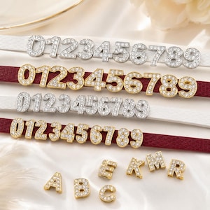 Peut inclure: Une collection de breloques chiffres en métal argenté et doré incrustées de strass transparents, disposées sur des bandes de cuir texturé blanc et bordeaux. Des lettres individuelles dorées, dont A, B, C, E, K, M, R, sont aussi présentes.