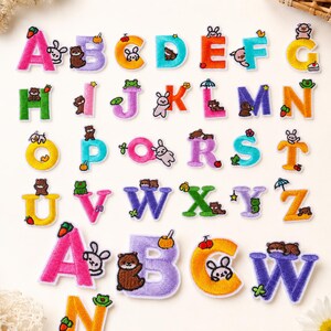 May include: A collection of embroidered alphabet patches featuring colourful letters paired with small animal and food illustrations. The set includes uppercase letters from A to Z, with additional larger letter patches at the bottom featuring bears.