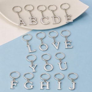 Puede incluir: Una colección de llaveros de metal en tono plateado, cada uno con un colgante de letra mayúscula unido a una anilla circular mediante una cadena corta. Se muestran las letras de la A a la J, incluyendo las palabras LOVE y YOU.