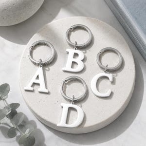 Könnte beinhalten: Vier silberfarbene Metallschlüsselanhänger mit den Großbuchstaben A, B, C und D. Jeder Buchstabe ist an einem runden Schlüsselring befestigt. Die Buchstaben haben eine glatte, polierte Oberfläche und sind auf einer runden Steinfläche angeordnet.