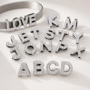 May include: A silver-toned mesh bracelet featuring the word LOVE spelled out in individual letter charms encrusted with clear crystals. Additional loose letter charms, also encrusted with clear crystals, are arranged below the bracelet.