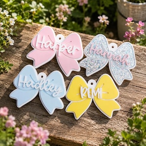May include: Four bow-shaped ornaments featuring personalized names. One is solid pink with the name Harper, one is silver glitter with the name Hannah, one is light blue with the name Hadley, and one is bright yellow with the name REMI. Each has a hole.