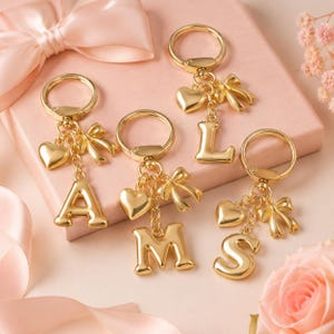 May include: Gold keyrings with letter charms, hearts, and bows. The letters A, L, M, and S are visible. The keyrings are displayed on a pink background with a pink ribbon and a pink gift box.