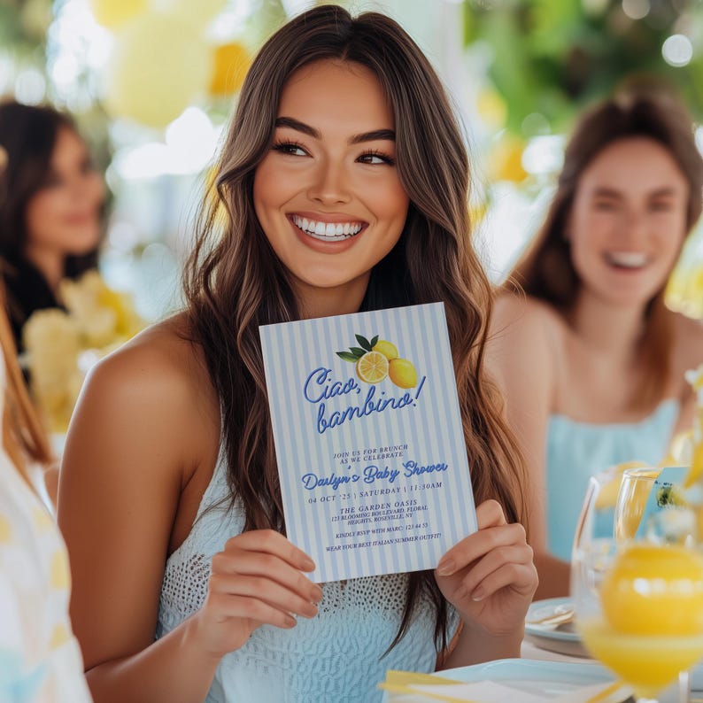 May include: A woman holds a blue and white baby shower invitation with the text "Ciao bambino!" and lemons. The invitation is for Darlyn's Baby Shower on October 21st at The Garden Oasis.