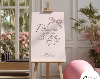 May include: A light pink sign on a wooden easel with the text "Pilates with the Birthday Girl" and a pink bow illustration. Below the text is "Nicole's 25th Birthday". A pink exercise ball is next to the easel.