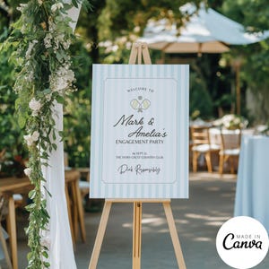 May include: An engagement party welcome sign on a wooden easel. The sign has a light blue and white striped background with text that reads "Welcome to Mark & Amelia's Engagement Party". The sign also includes the date and location.