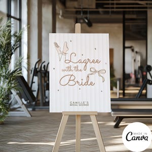 May include: A wooden easel displays a sign with the text "Lagree with the Bride" in a decorative font, along with a bow illustration. The sign is for Camille's bridal shower. The background is a gym.