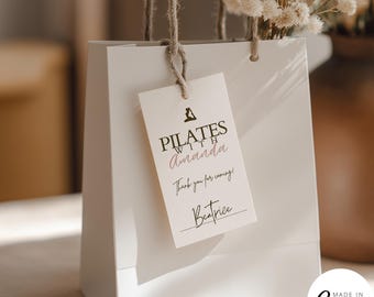Puede incluir: Una bolsa de regalo blanca con una etiqueta que dice "PILATES with Amanda". La etiqueta también dice "Thank you for coming! Beatrice". La bolsa tiene un asa de cuerda marrón y está decorada con flores secas.