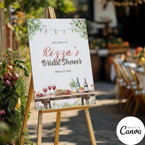 May include: A wooden easel displays a sign that reads "Welcome to Rizza's Bridal Shower, March 27, 2025." The sign features watercolor illustrations of wine glasses, bottles, and food. Greenery and string lights decorate the top.