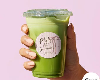 May include: A clear plastic cup filled with a vibrant green beverage, topped with a clear lid. A round sticker on the cup reads "Pilates with Samantha" with a pink bow illustration. The background is a soft pink.