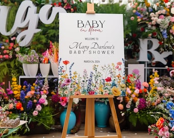 Könnte beinhalten: Ein weißes Schild mit einem Blumenmuster und dem Text "Baby in Bloom" in rosa Schrift. Das Schild zeigt auch "Willkommen zur Babyparty von Mary Darlene" und "March 24, 2024". Das Schild steht auf einem Holzstaffelei.