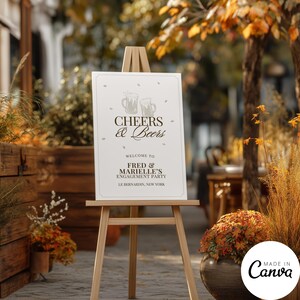 May include: A white rectangular sign on a wooden easel featuring the text Cheers & Beers, Welcome to Fred & Marielle's Engagement Party, and Le Bernardin, New York, with two beer mug illustrations and small confetti-like accents in dark gray.