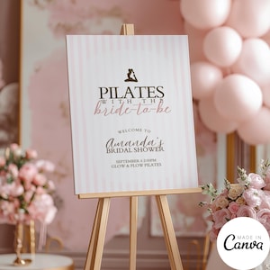 May include: A sign on a wooden easel with the text "PILATES with the bride-to-be" and "Amanda's Bridal Shower". The sign has a pink and white striped background and is surrounded by pink balloons and flowers. The sign says "September 6 2:00PM Glow & Flow Pilates".