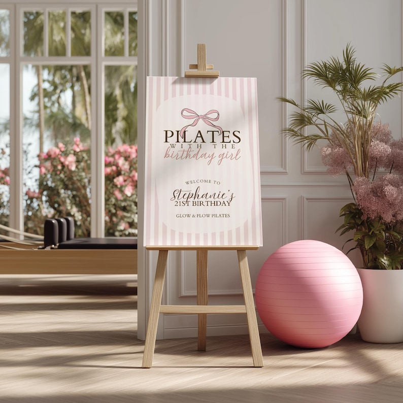 Peut inclure: Un chevalet en bois pr&eacute;sente une affiche avec le texte "Pilates with the birthday girl". L'affiche a un fond &agrave; rayures roses et blanches et un motif de n&oelig;ud rose. Un ballon d'exercice rose et des plantes en pot sont en arri&egrave;re-plan.