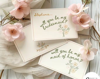 May include: Two cards with floral illustrations and the text "Stephanie... will you be my bridesmaid?" and "will you be my maid of honor?"  The cards are on a white background with pink flowers.