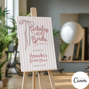 May include: A pink and white striped welcome sign on a wooden easel. The sign reads "Pilates with the Bride" and welcomes guests to Amanda's Bridal Shower on September 6, 2025. A decorative bow is at the top.