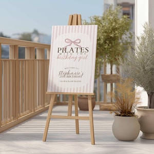 Peut inclure: Un chevalet en bois supportant une affiche avec le texte "Pilates with the birthday girl". L'affiche pr&eacute;sente un fond &agrave; rayures roses et blanches et un motif de n&oelig;ud. Elle souhaite la bienvenue &agrave; l'anniversaire de St&eacute;phanie.