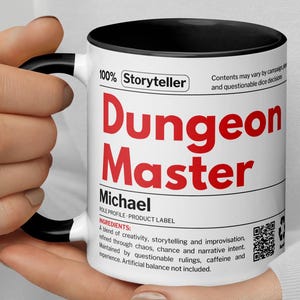 Puede incluir: Una taza de cerámica blanca con interior y asa negros. La taza presenta las palabras "Dungeon Master" en rojo, junto con texto adicional. La taza está sostenida por una persona.